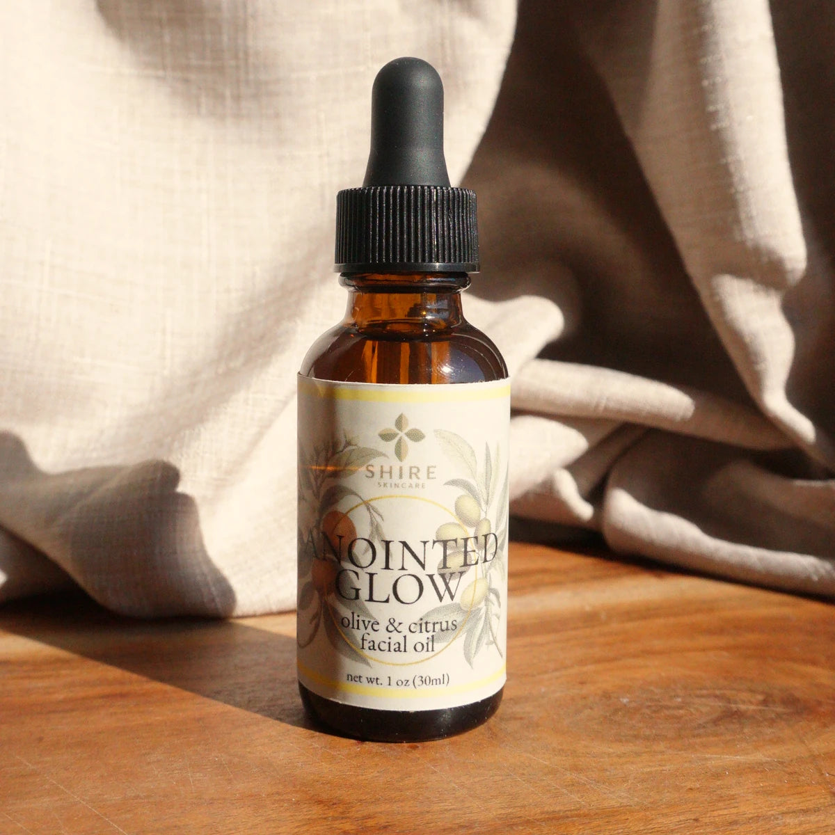 Bottle of biblical skincare product called "Anointed Glow" Olive & Citrus Facial Oil on a wooden surface with beige fabric in the background