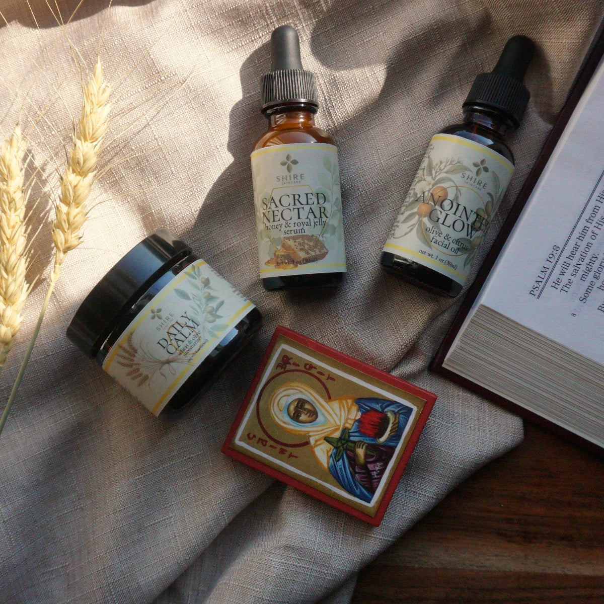 Three bottles and a jar labeled 'Sacred Nectar', 'Anointed Glow', and 'Daily Calm' on a textured surface with a Bible and icon of St Brigit.