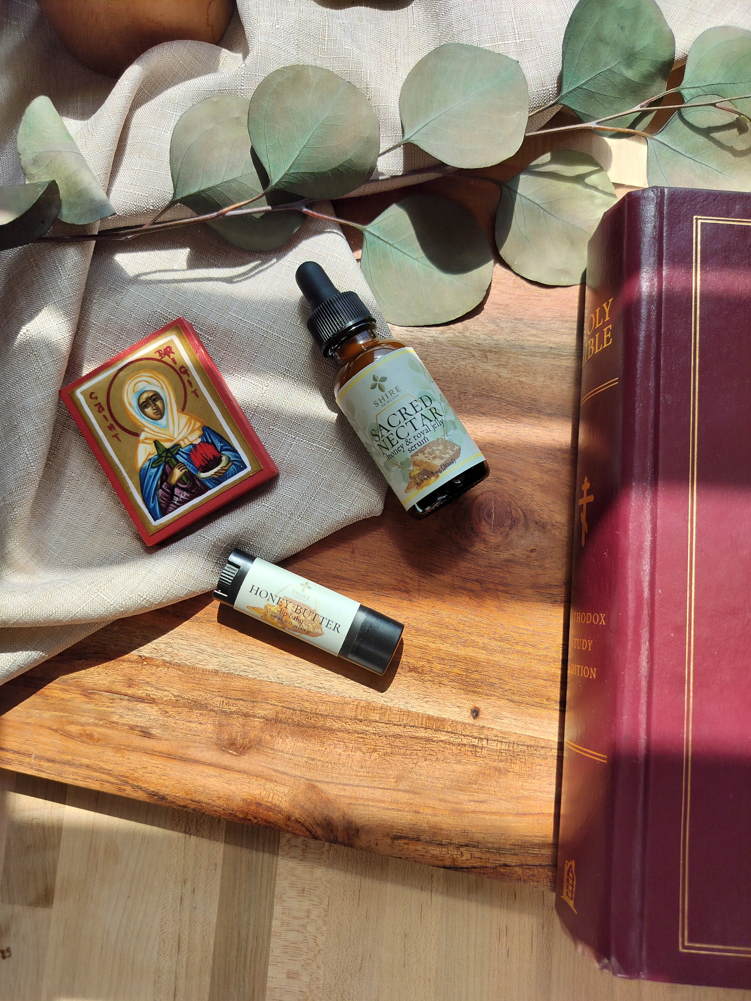 Bottle of 'Sacred Nectar' serum, and Honey Butter Lip Balm,  a Holy Bible, and an icon of Saint Brigit on a wooden surface with eucalyptus leaves.