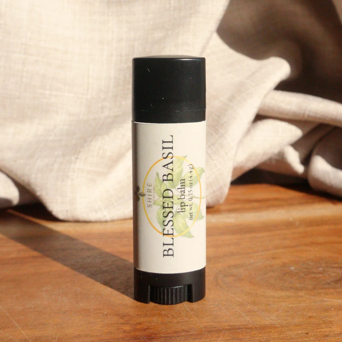 Lip balm stick labeled 'Blessed Basil' on a wooden surface with a soft fabric background.