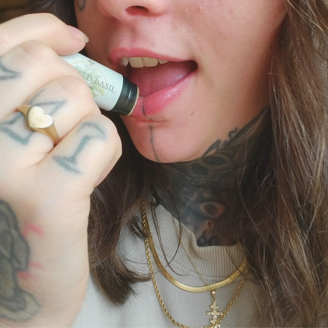 Person applying a lip balm stick called "Blessed Basil" with visible tattoos and jewelry.