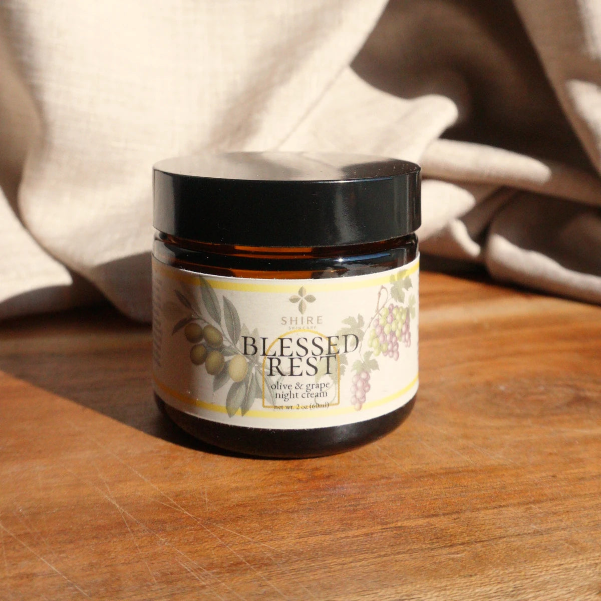 Jar of 'Blessed Rest' olive and grape night cream on a wooden surface with a soft fabric background