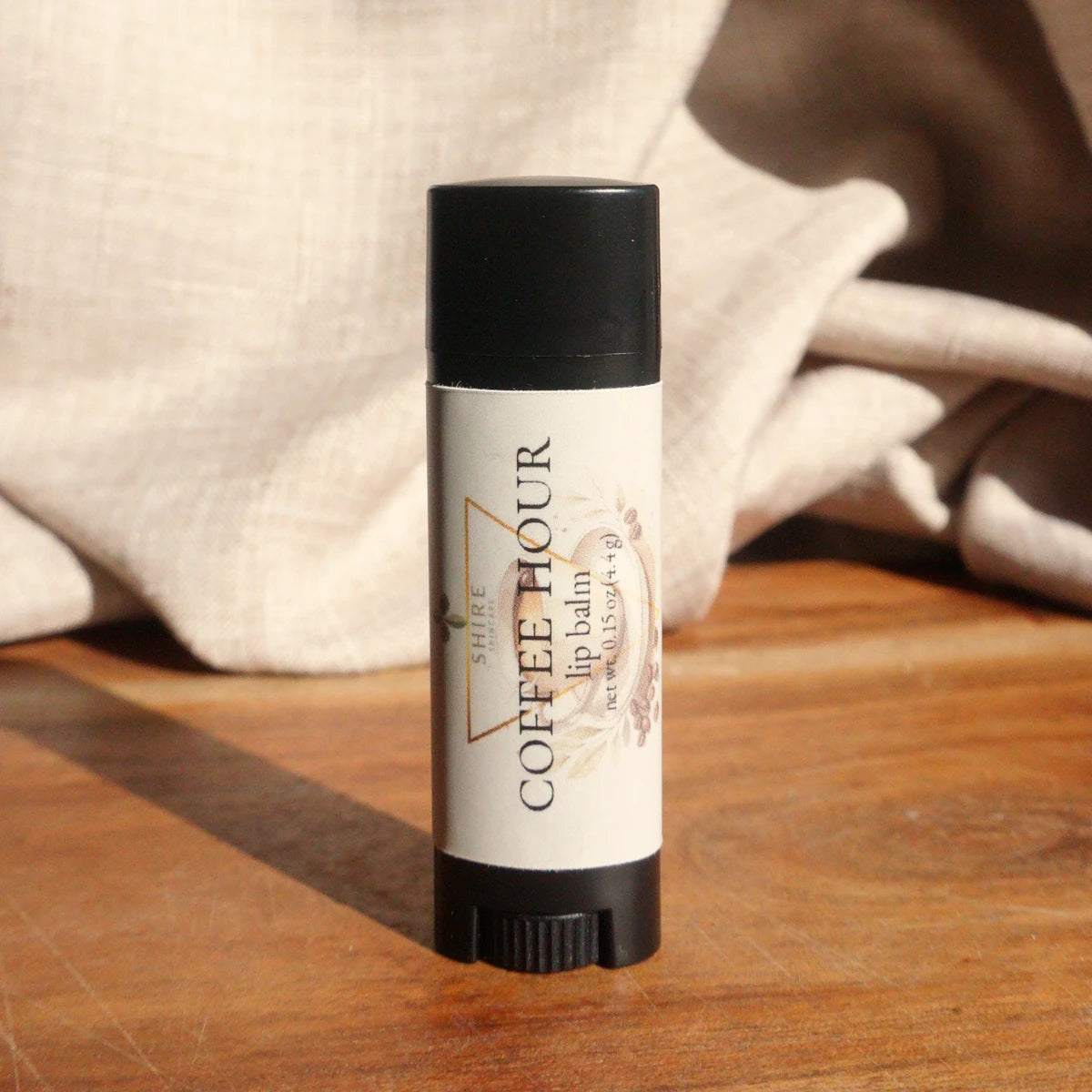 Lip balm tube labeled 'Coffee Hour' on a wooden surface with a soft fabric background