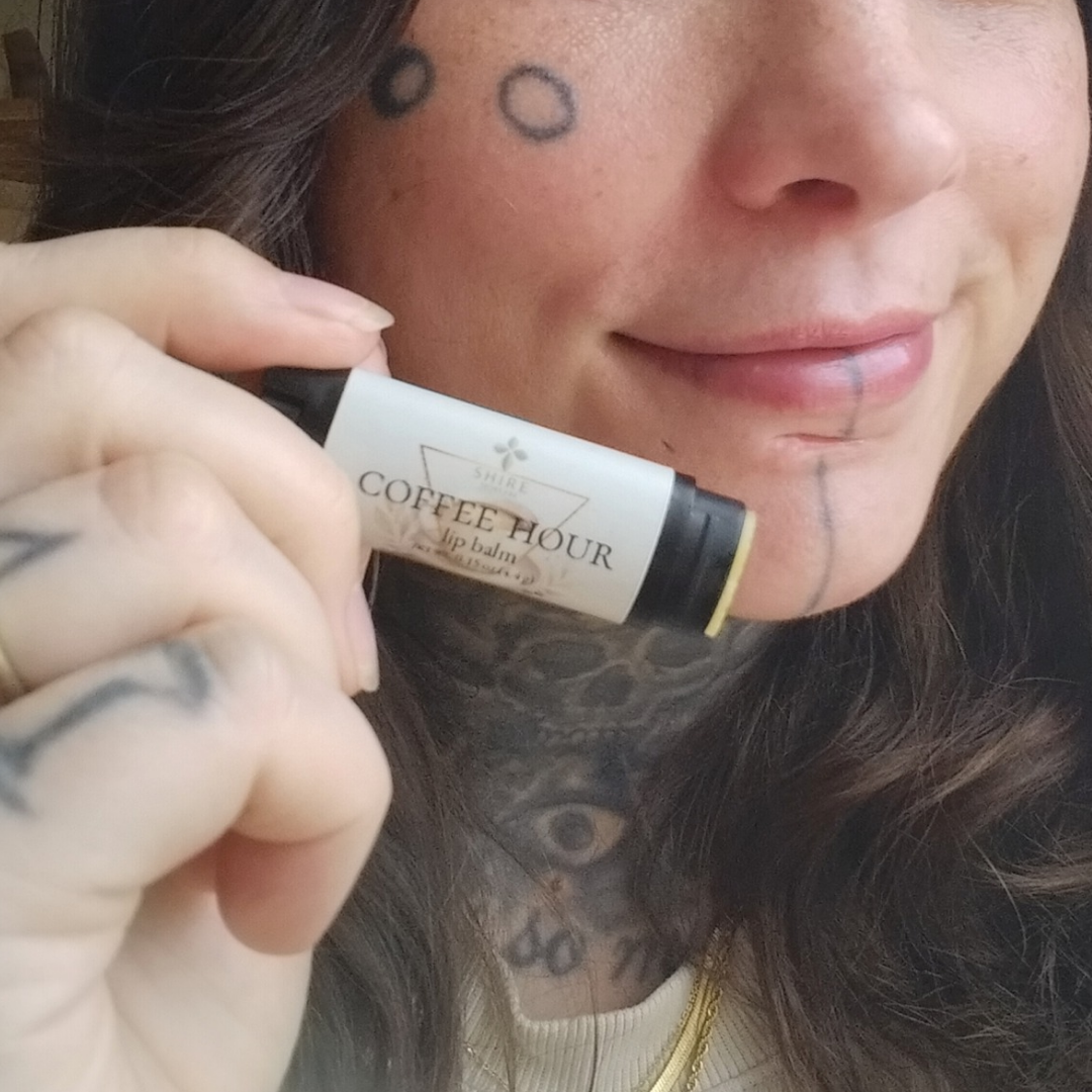 Person holding a lip balm labeled 'Coffee Hour' with tattoos visible on face and neck.