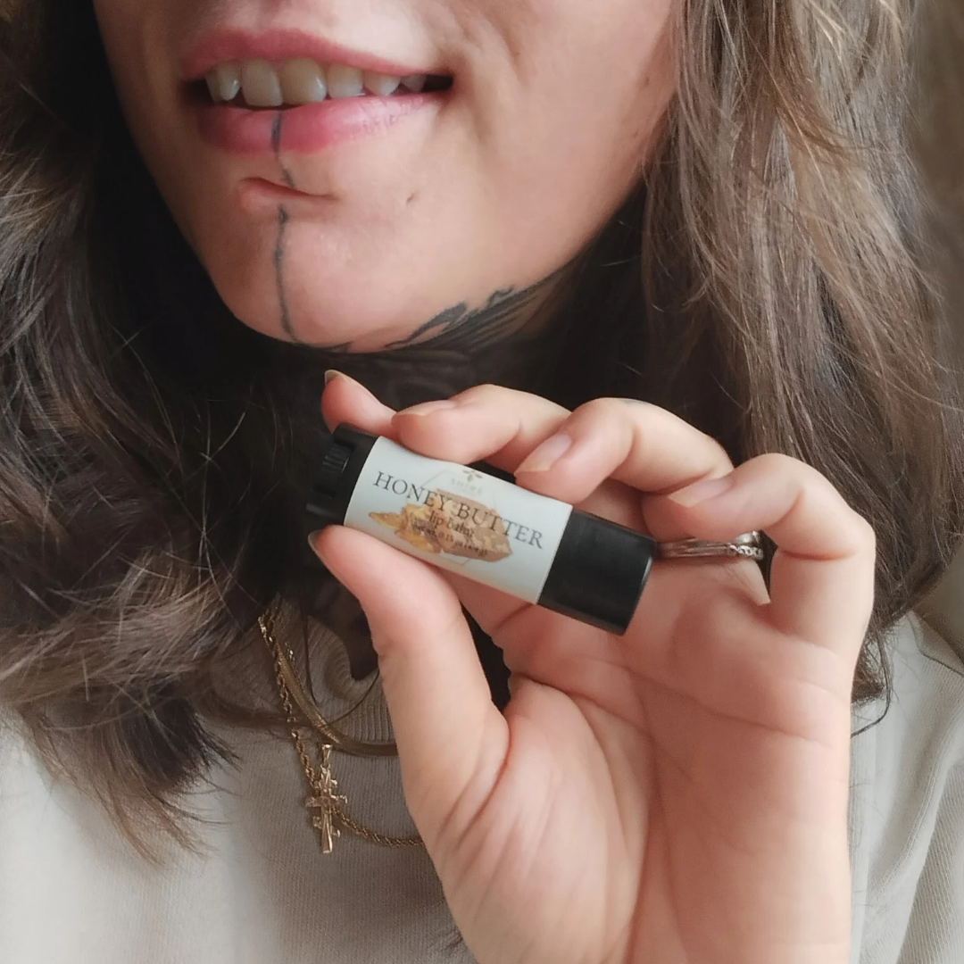 Person wearing a cross holding a lip balm labeled 'Honey Butter' close to their mouth.