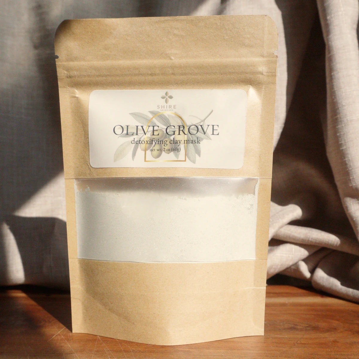 Kraft paper packaging of Olive Grove clay mask on a wooden surface with a neutral background