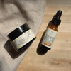 Two skincare products on a wooden surface: a jar labeled 'Daily Calm' and a dropper bottle labeled 'Sacred Nectar'.