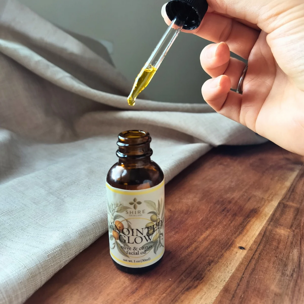 Hand holding a dropper with a small amount of golden liquid above a bottle labeled 'Anointed Glow' facial oil on a wooden surface.
