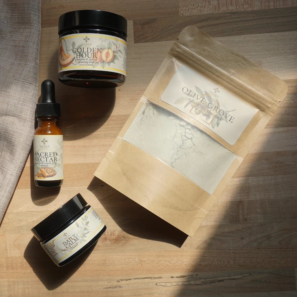 Assorted natural skincare products including moisturizer jars, a serum dropper bottle, and a clay mask in a paper bag on a wooden surface.