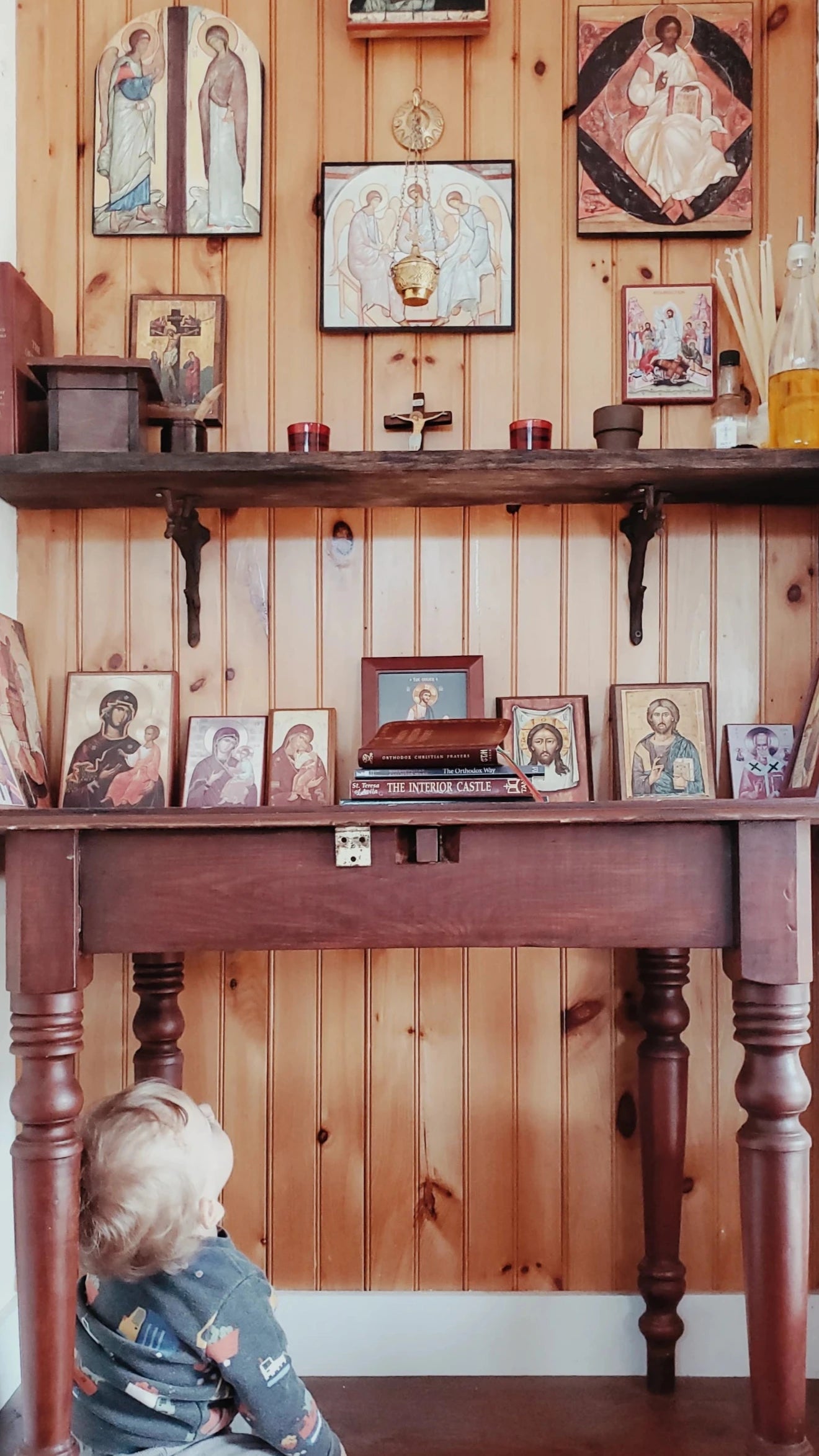 Child sitting on the floor looking at a wooden shelf with various Orthodox icons  in a prayer corner.